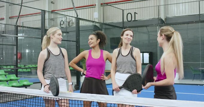 Female friends holding rackets, ball at net laughing as fuchsia top speaking on padel court