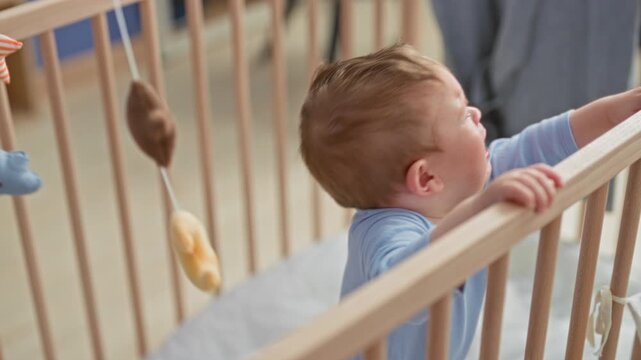Toddler crying and reaching with open hand while gripping wooden crib rail in nursery; distress seeking comfort.