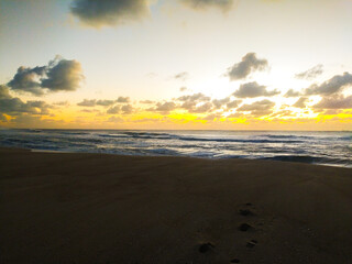 Obraz premium Experience the serene beauty of a golden ocean sunrise. This cinematic coastal landscape features footprints in the sand, rolling waves, and dramatic clouds, perfect for travel concepts.