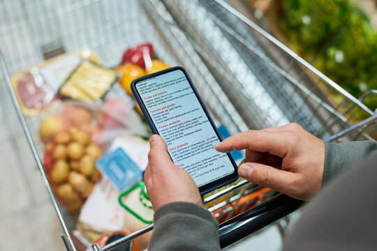 Shopper checking grocery list on smartphone while pushing cart through supermarket, showing retail planning and budgeting. Useful for food shopping, consumer app, retail marketing