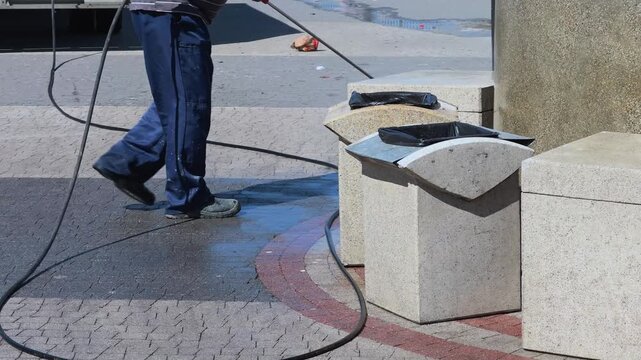 Anonymous male municipal utility worker in uniform cleaning public stone trash bins and street pavement using commercial high pressure water washer on bright sunny summer day medium close up.