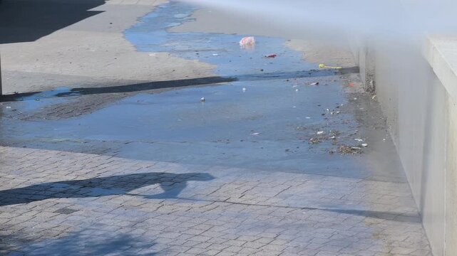 Anonymous municipal groundskeeper legs washing stone parapet wall and dirty cobblestone promenade pavement with heavy duty high pressure water jet spray under bright summer sunlight authentic shot