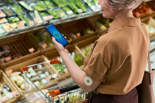 Adult woman with diabetes checking glucose app while shopping in supermarket produce aisle. Useful for diabetes management, healthy grocery choice marketing