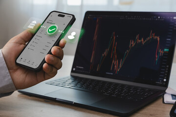 Hand holding smartphone with online banking interface beside laptop displaying financial trading...