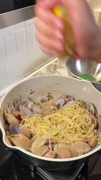 Asian female influencer cooking vlog video using smartphone showing spaghetti alle vongole recipe with fresh clams parsley and lemon juice in modern kitchen atmosphere squeezing lemon over clam pasta