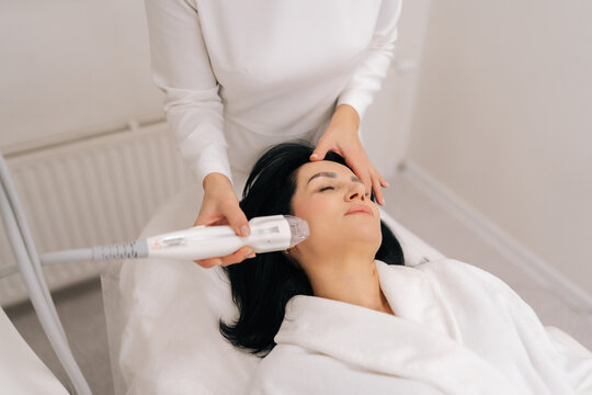 Top view of relaxed adult woman receiving innovative rf microneedling facelift procedure from skilled esthetician, modern hardware cosmetology method for skin tightening and wrinkle reduction.