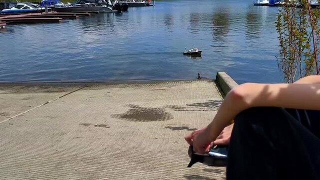 A man controls a model ship by the water against the background of a yacht marina, a calm pond and a summer scene of ship modeling.
