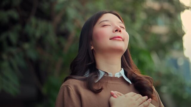 Happy Asian woman closed eyes holding hands on her chest, breathing fresh air in the green garden park. Peaceful female relaxing outdoors, feeling grateful, relieved, and mindful for mental health
