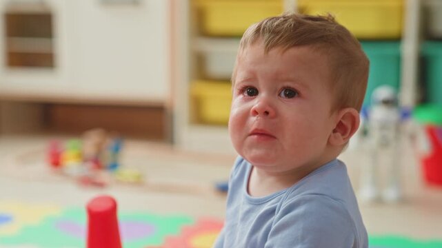 Boy crying with tears on his face in a colorful playroom among scattered toys, foam mat and a red bucket; sadness.