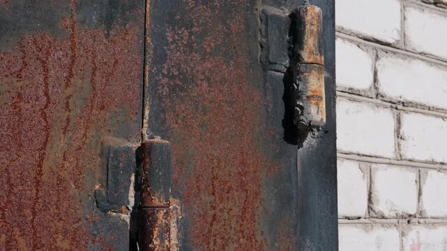 Close pan along a heavily rusted metal door with welded iron hinges and peeling paint next to a white brick wall. This represents an aging urban infrastructure concept.