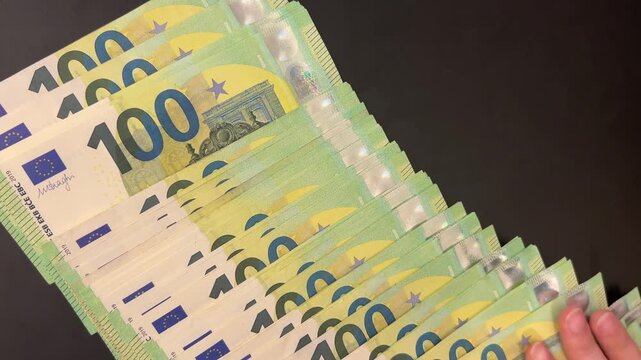 Hands spreading stack of 100 euro bills across dark table. 100 euro banknotes being spread out on black surface, close-up