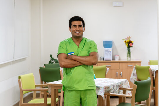 Male nurse wearing green scrubs standing with crossed arms, smiling at camera in a nursing home dining area