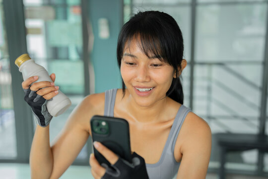 Sweaty young Asian woman in sportswear drinking a protein shake from a bottle and using her smartphone while resting after a workout at the gym.