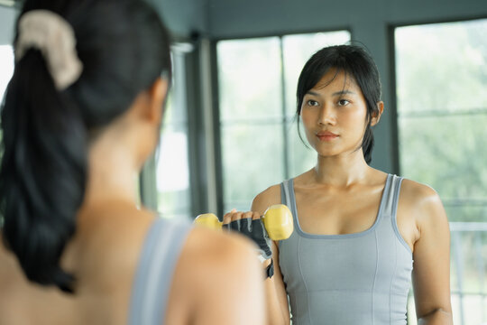 Asian woman in gray sportswear holds yellow dumbbells at her shoulders while looking at her reflection in a gym mirror.