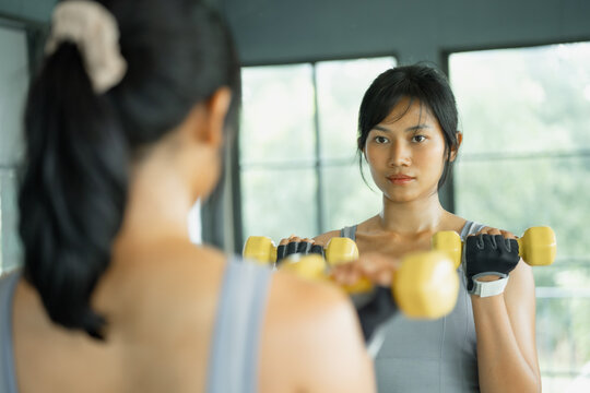 Asian woman in gray sportswear extends her arms in a lateral raise with yellow dumbbells during a strength workout in a gym.