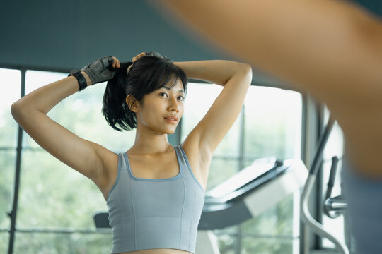 Asian woman in gray sportswear and workout gloves ties her ponytail as she prepares to train by bright gym windows.