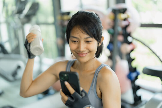 Asian woman in gray sportswear smiles at her smartphone while holding a shaker bottle of protein drink in a bright gym.