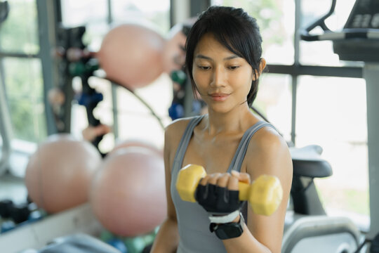 Asian woman in gray sportswear smiles at her phone while holding a shaker bottle of protein drink during a break in a bright gym.