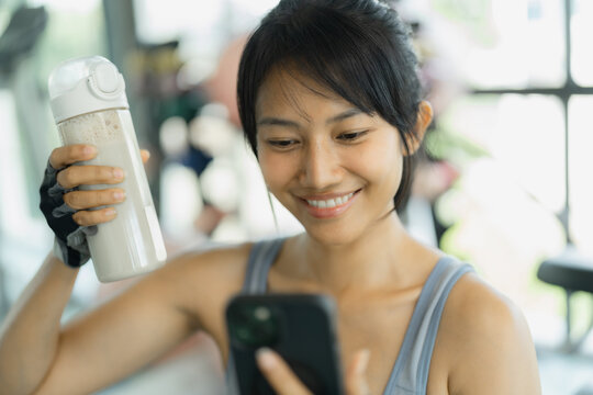 Asian woman in gray sportswear smiles at her smartphone while holding a shaker bottle of protein drink among gym equipment.