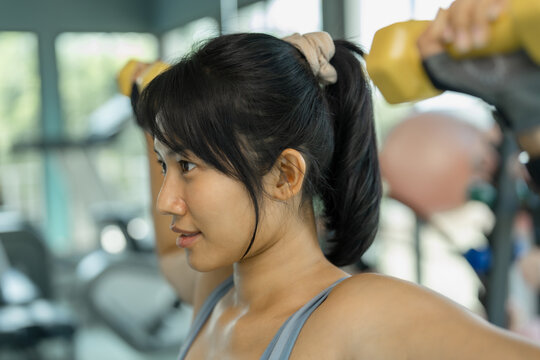 Close-up profile of an Asian woman in gray sportswear lifting a yellow dumbbell during a focused workout in a bright gym.
