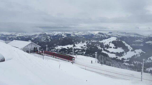 Red cogwheel train at Rigi Kulm, the summit station of Mount Rigi in central Switzerland. Panoramic winter landscape featuring snow-covered Alpine peaks, ski slopes and dramatic mountain scenery.