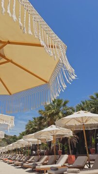 Beautiful 4K low-angle shot of white fringed macrame umbrellas at a luxury beach resort. Features an organized row of sunbeds, tropical palm trees, and a clear blue sky.