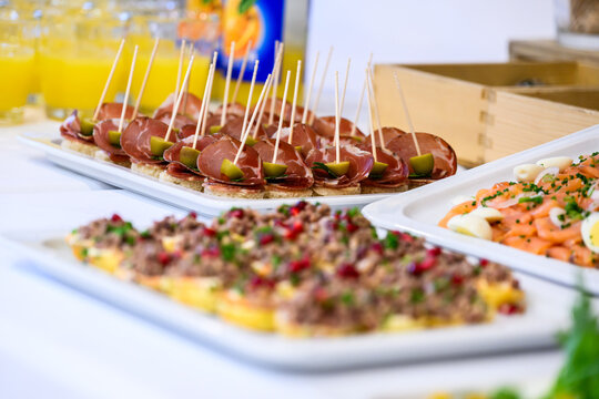 A vibrant catering spreadsheet displaying toothpick skewers of cured ham and green olives on small bread bases. The background features fresh juices and adjacent buffet platters, all presented with a