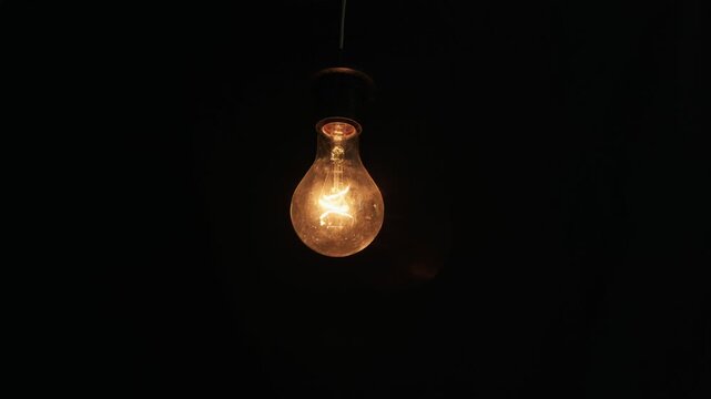A single vintage incandescent light bulb with a glowing filament hangs suspended against a pitch-black background. Concept of bright ideas, electrical energy, and innovation.