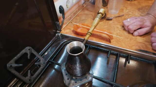 A traditional engraved copper cezve brews fresh morning coffee over a lit gas stove burner. A man hands rest patiently on the warm kitchen counter in the blurred background.