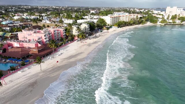 sandy bay of St Lawrence Gap beach with luxury resorts, lush trees and turquoise sea waves breaking on shore in Barbados drone aerial footage