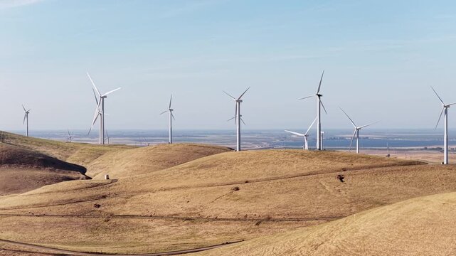 Wind turbines generating clean, renewable energy spinning over the rolling, dry hills of the Altamont Pass Wind Farm in Livermore, California, under a clear blue sky on a sunny day