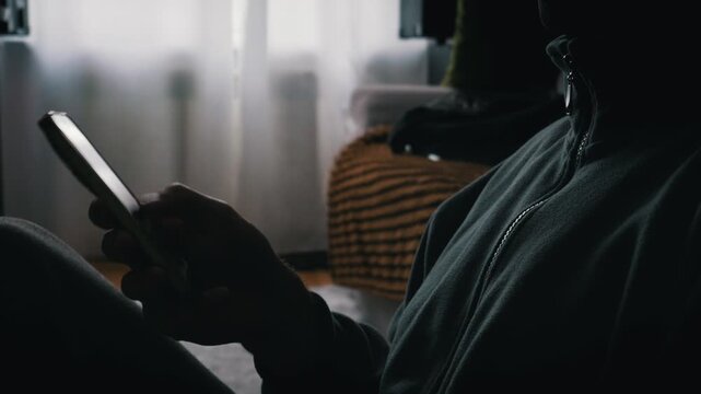 A person in a dark green fleece jacket scrolls on a smartphone in a dimly lit room. The screen glows brightly against the shadows, creating a quiet and focused atmosphere.