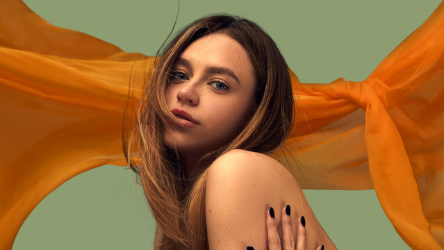 Beauty portrait with orange sheer fabric and hand on bare shoulder. Concept of skincare advertising, fashion editorials, cosmetics branding, beauty campaigns, sensual studio mood.