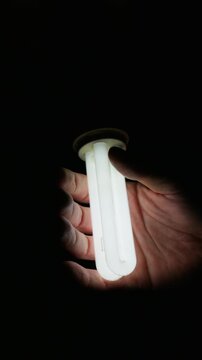 Person hand holds a brightly glowing compact fluorescent lamp in total darkness. The soft white light illuminates the fingers and palm, creating a strong contrast against the black background.