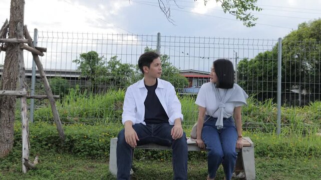 Happy young Asian couple sitting on a bench together, talking and smiling during a romantic date in a public park