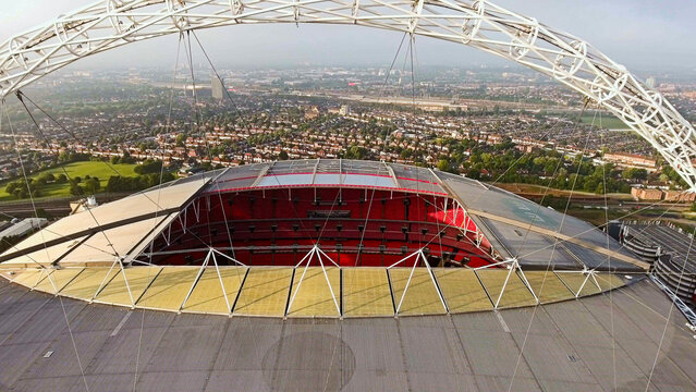 Aerial drone close-up of Wembley Stadium arch with steel lattice truss and cable stays framing the red seating bowl, Wembley, London, England