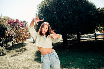 Smiling young woman posing playfully in a park wearing a cropped sweatshirt and jeans on a sunny...