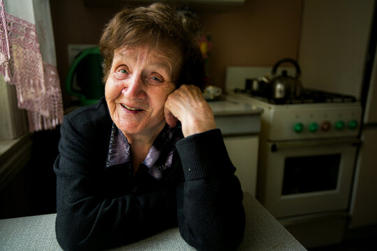 Smiling elderly woman sitting at a kitchen table near a window in a modest home, looking warmly toward the camera.