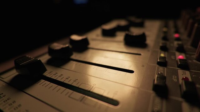 Finger pushes down a fader on a professional audio mixer console to reduce the sound volume. Deep blue atmospheric lighting illuminates the studio equipment and highlights the sliders.