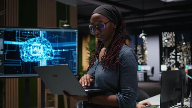 Female technician in office using laptop, working to fix bugs in artificial intelligence system. Woman troubleshooting neural network errors with notebook in tech agency workplace.