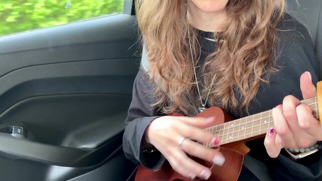 Slow motion close up of teenage girl playing ukulele while riding in car during family road trip. Young musician practicing instrument and enjoying creative leisure activity during travel.