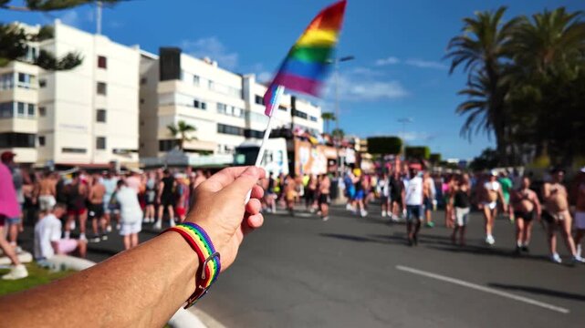 Person's hand waving a small rainbow flag with a rainbow bracelet during a sunny outdoor gay pride parade, with a crowd of people walking and celebrating on the street in the background