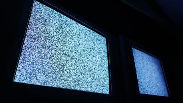 Two glowing Cathode Ray Tube television screens display static white noise in a dark room. The loss of broadcast signal conveys an eerie retro technology concept, suggesting a bygone era of