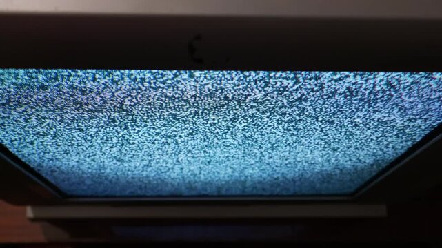 Analog television screen displays a continuous white noise static pattern. Erratic black and white pixels create a strong visual texture. A dark plastic bezel frames the glowing interference