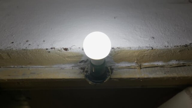 A bright LED light bulb glows in a socket mounted on a cracked wall with peeling paint in a dim room. Concept of illumination, urban decay, outdated interiors, and hope in dark spaces.