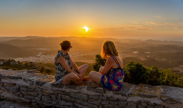 Two young female friends in summer dresses sitting on a stone wall watching a spectacular sunset during a solar eclipse from the Monte Toro viewpoint in Menorca, Spain.