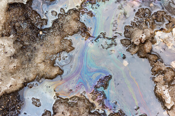 Fototapeta premium Iridescent oil slick swirling in a melting snow puddle on an asphalt road surface during spring thaw, creating colorful chemical patterns mixed with slush and dirty winter grit