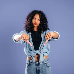 Young woman with curly hair wearing a denim jacket and black shirt gestures with thumbs down against a solid purple background in a studio setting © standret