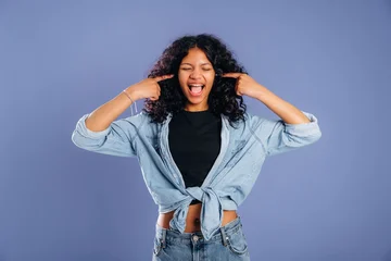 Young woman with curly hair wearing denim shirt and black top playfully covers her ears while smiling against a solid purple background © standret