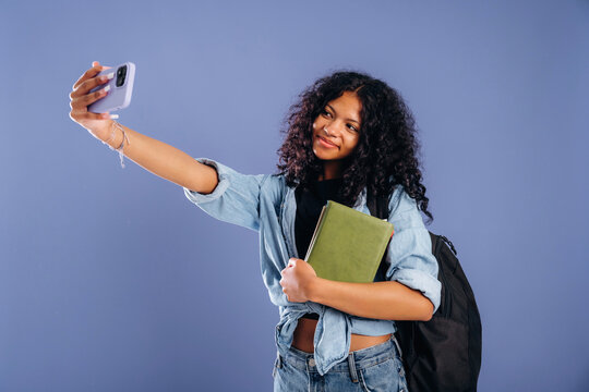 Young African American girl with curly hair takes a selfie while holding a green notebook and wearing a black backpack against a blue background